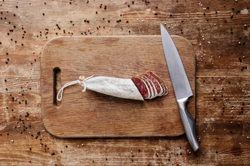 top view of cutting board with steel knife and tasty sliced salami on wooden table with scattered...