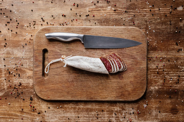 top view of wooden cutting board with steel knife and sliced salami on table with scattered spices