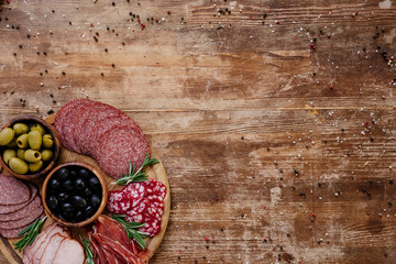 top view of cutting board with olives, breadsticks, delicious prosciutto, salami and herbs on wooden table with scattered spices