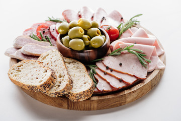 round cutting board with delicious salami, ham, olives in bowl, bread and herbs on white table