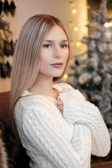 Young beautiful blonde girl in a warm white knitted sweater on the background of a homely cozy room with Christmas garlands.