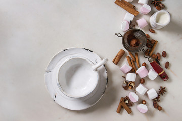Ingredients for hot chocolate. White pink big marshmallow, cocoa beans, cocoa powder, cinnamon, cream, chopped chocolate, nuts, empty ceramic cup over white marble background. Flat lay, space.