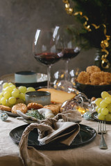 Home Christmas or New year dinner table set with wine, cheese plate, appetizers, lightning garland, xmas decoration and empty plate with cloth napkin.