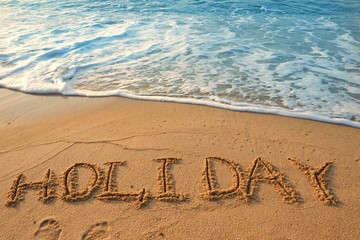 Concept HOLIDAY handwriting on sand beach with waves and blue ocean.