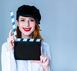 Young redhead woman in hat holding movie clapboard on grey background