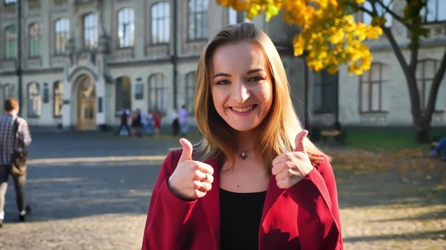 Young beautiful female student shows thumbs up, happy and positive, being outside the university in a sunny day
