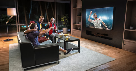 Group of fans are watching a soccer moment on the TV and celebrating a goal, sitting on the couch in the living room.