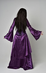 full length portrait of beautiful girl with long black hair,   wearing purple fantasy medieval gown. standing pose  with back to the camera on grey studio background.