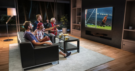 Group of fans are watching a soccer moment on the TV and celebrating a goal, sitting on the couch in the living room.