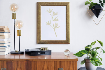Retro and minimalistic interior with gold mock up poster frame on the vintage brown shelf, hanging plant in design pot, books, platns, table lamp and box.  