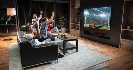 Group of students are watching a soccer moment on the TV and celebrating a goal, sitting on the couch in the living room.