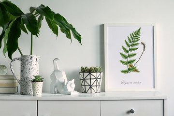 Scandinavian stylish cupboard with mock up poster frame, cat figure, tropical leaf, accessories, books, cacti. White background wall.