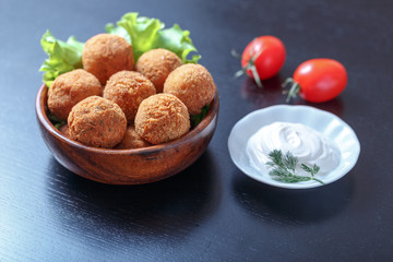 Falafel lies in a wooden plate. On the table lie tomatoes, cucumbers, lettuce, dill, lemon, sour cream.