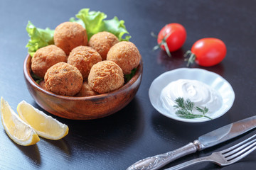 Falafel lies in a wooden plate. On the table lie tomatoes, cucumbers, lettuce, dill, lemon, sour cream.