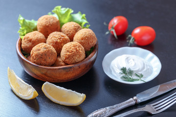 Falafel lies in a wooden plate. On the table lie tomatoes, cucumbers, lettuce, dill, lemon, sour cream.