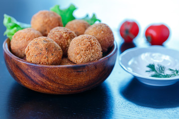 Falafel lies in a wooden plate. On the table lie tomatoes, cucumbers, lettuce, dill, lemon, sour cream.