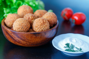 Falafel lies in a wooden plate. On the table lie tomatoes, cucumbers, lettuce, dill, lemon, sour cream.