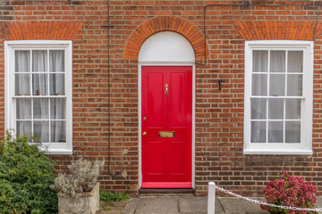 Fototapeta na wymiar Typical english house facade with red door, two white windows and bricks wall viewed from outdoors.