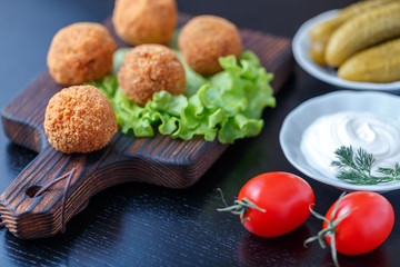 Falafel lies on a wooden cutting board. On the table lie tomatoes, cucumbers, lettuce, dill, lemon, sour cream.