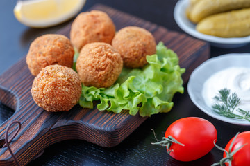 Falafel lies on a wooden cutting board. On the table lie tomatoes, cucumbers, lettuce, dill, lemon, sour cream.