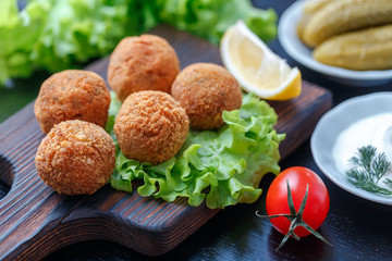 Falafel lies on a wooden cutting board. On the table lie tomatoes, cucumbers, lettuce, dill, lemon, sour cream.
