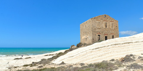 Naklejka premium Woman on empty white beach with old ruins of abandoned stone house on the rocks and summer blue sky and sea in background in Sicily Italy