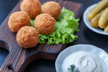 Falafel lies on a wooden cutting board. On the table lie tomatoes, cucumbers, lettuce, salad, dill, lemon, sour cream.
