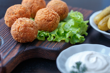 Falafel lies on a wooden cutting board. On the table lie tomatoes, cucumbers, lettuce, salad, dill, lemon, sour cream.