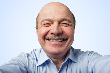 Elderly man with a mustache holding a smartphone and makes selfie, smiling broadly on blue...