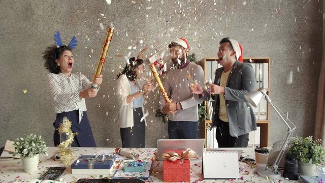 Multiethnic group of young male and female office workers blowing firecrackers, drinking champagne and laughing happily while having corporate Christmas party