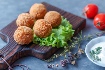 Falafel lies on a wooden cutting board. On the table lie tomatoes, cucumbers, lettuce, salad, dill, lemon, sour cream.