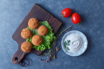 Falafel lies on a wooden cutting board. On the table lie tomatoes, cucumbers, lettuce, salad, dill, lemon, sour cream.
