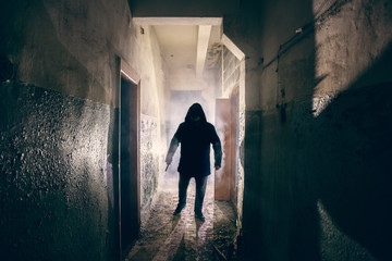 Dark silhouette of strange danger man with knife in hand in hood in back light with smoke or fog in scary grunge corridor or tunnel - obrazy, fototapety, plakaty