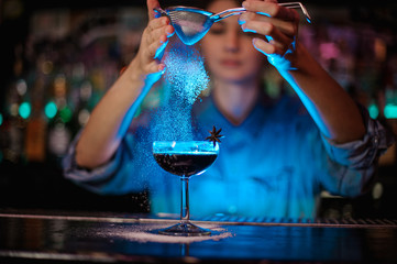 Girl adding to a brown cocktail a sugar powder through the strainer in the blue light