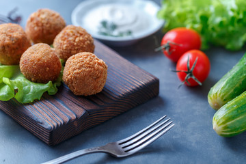Falafel lies on a wooden cutting board. On the table lie tomatoes, cucumbers, lettuce, salad, dill, lemon, sour cream.