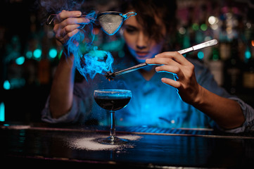 Female bartender pouring on the brown cocktail and on a flamed badian on tweezers a powdered sugar...