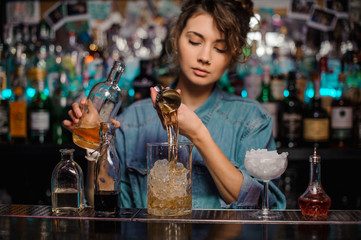 Bartender girl pouring to the measuring glass cup with ice cubes an alcoholic drink from jigger