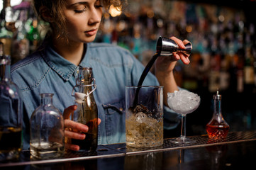 Female bartender pouring to the measuring glass cup with ice cubes a brown alcoholic drink from steel jigger