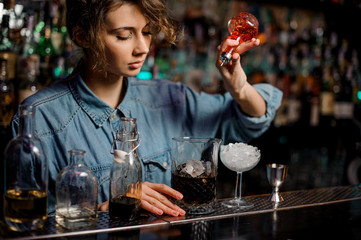 Bartender girl pouring to the measuring cup with ice cubes a red bitter from the glass bottle