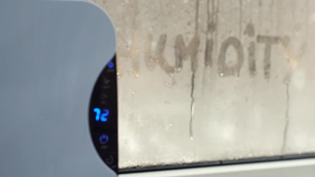 Woman turns dehumidifier on using touch panel by wet window. Humidity indicator shows high level of dampness in flat