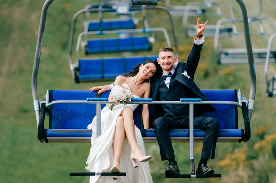 Happy wedding couple riding cableway from mountain in summer. Smiling cheerful bride in white elegant dress lying on grrom shoulder in cable car. Funny groom showing devil horns gesture with hand.