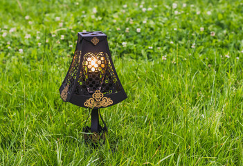 A beautiful black openwork metal retro lamp with a gold ornament and with a yellow burning lamp on the background of green grass