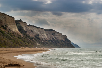 A view after a summer storm on a sandy beach located beneath tall rocks with greenery and a cloudy sky with penetrating sun rays. Gardenos beach, Corfu island.