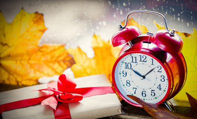 Red vintage alarm clock and gift box with maple leaves on rainy background