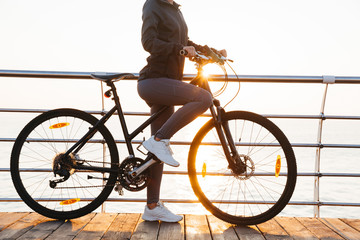 Photo of slim woman riding bicycle on boardwalk, during sunrise over sea