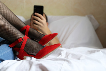 Fototapeta premium Couple in bed, sexual issues in family and mobile gadgets. Man lying with smartphone in hand near girl in fishnet stockings and red shoes on high heels, concept of online or gaming addiction