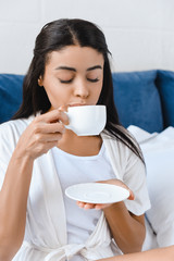 attractive mixed race girl in white robe drinking coffee in bed in morning with closed eyes