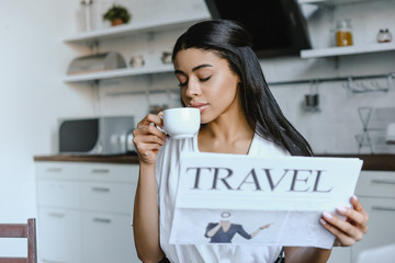 beautiful mixed race girl in white robe drinking coffee and holding travel newspaper in morning in kitchen