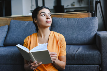 beautiful mixed race girl in orange shirt holding book in living room and looking away - Powered by Adobe