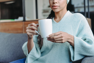 cropped image of mixed race girl in turquoise sweater and headband sitting on sofa and holding cup of tea in living room